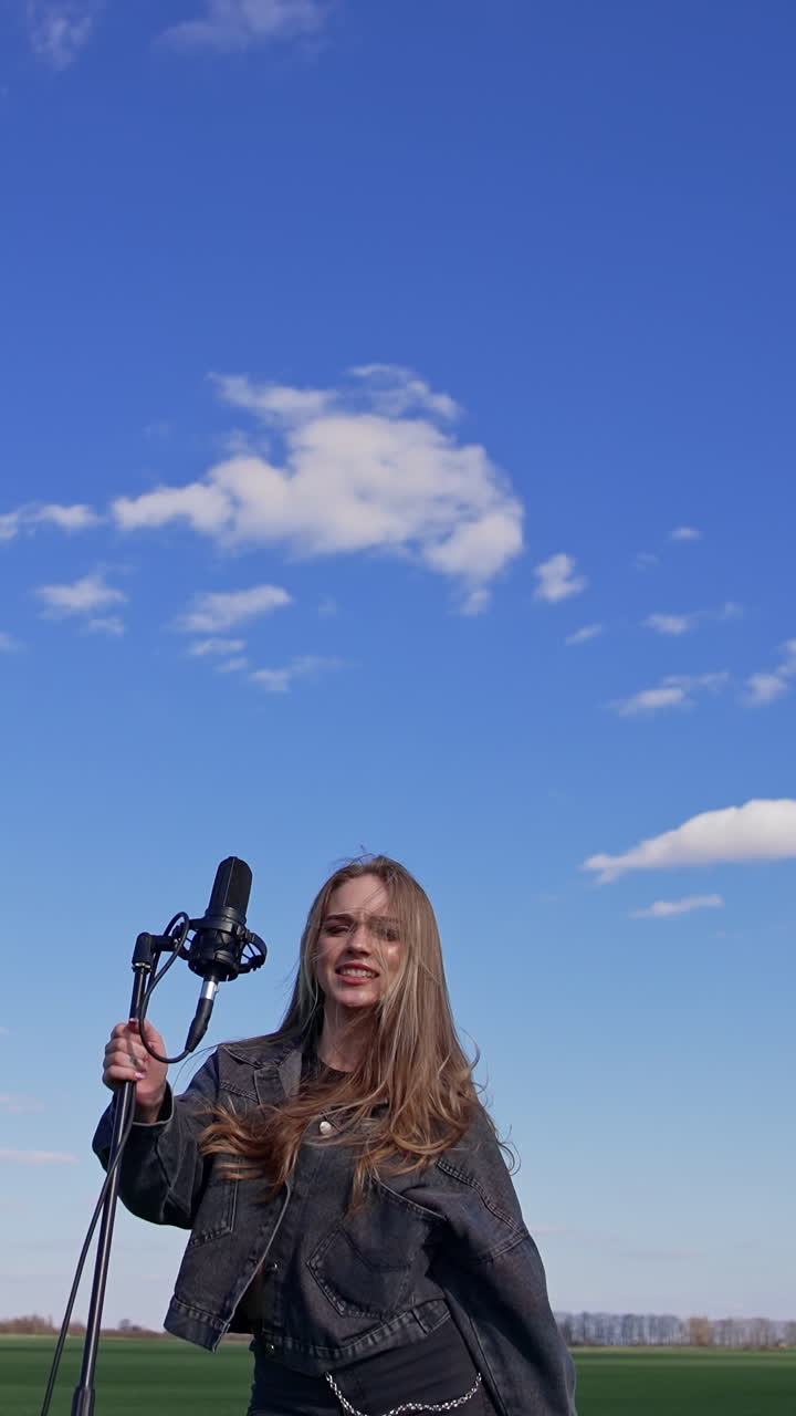Beautiful young singer dancing with a microphone on a field. Pretty female soloist singing and jumping cheerfully outdoors. Slow motion. Vertical video