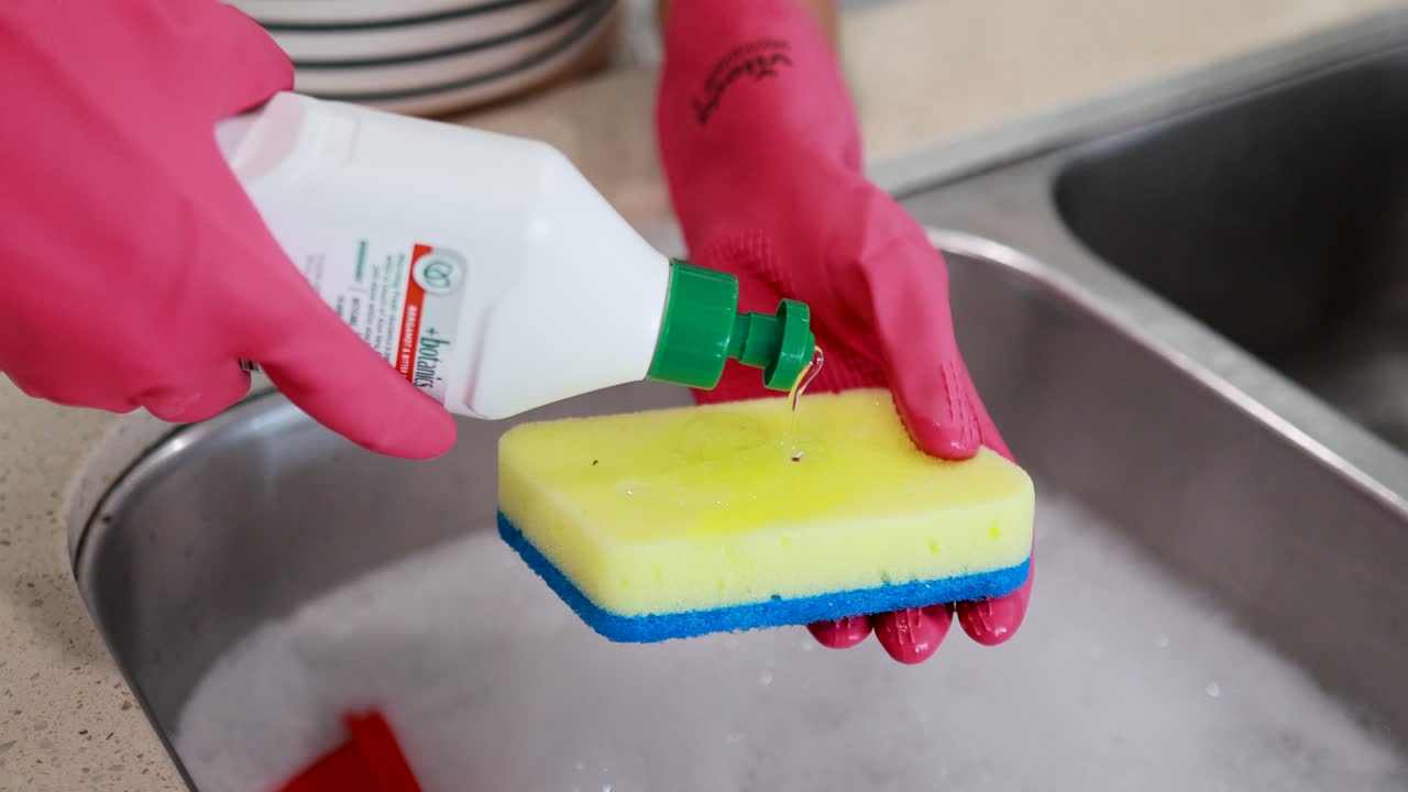 Hands in gloves applying dish soap to a sponge over a sink. Bright lighting, close-up view, domestic setting