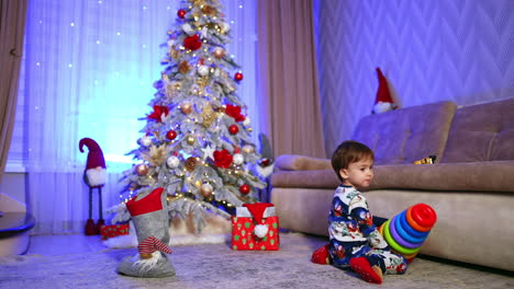 little caucasian toddler playing in the room decorated for christmas holidays. sweet kid holds a colorful toy pyramid.