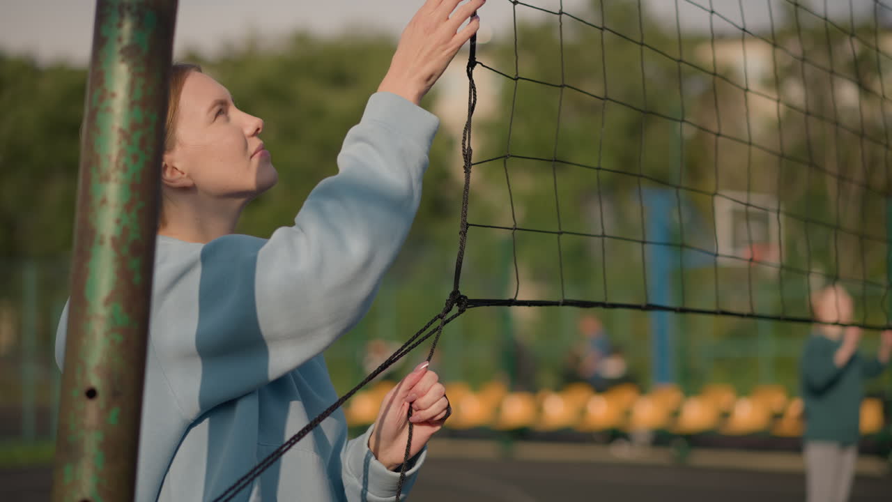 Lady holding volleyball net with two women playing volleyball in the background, other people in distant blurred background on sunny outdoor sports field