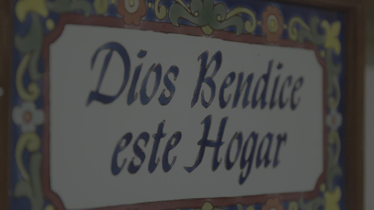 Close-up shot of a colorful, hand-painted ceramic sign with the Spanish blessing 'Dios Bendice este Hogar'. Soft lighting and intricate floral details create a warm, inviting atmosphere