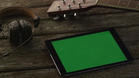 tablet pc with green screen laying on wooden table next to guitar and headphones. causal lifestyle