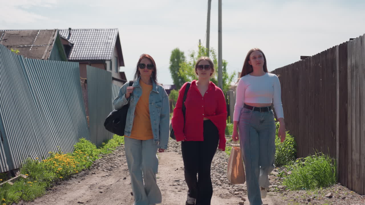 Three women walking side by side on narrow rocky village road surrounded by fences and houses, carrying bags and enjoying bright sunny day, dressed casually in cheerful mood under clear sky
