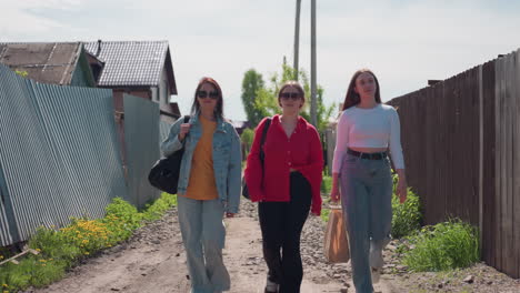 three women walking side by side on narrow rocky village road surrounded by fences and houses, carrying bags and enjoying bright sunny day, dressed casually in cheerful mood under clear sky