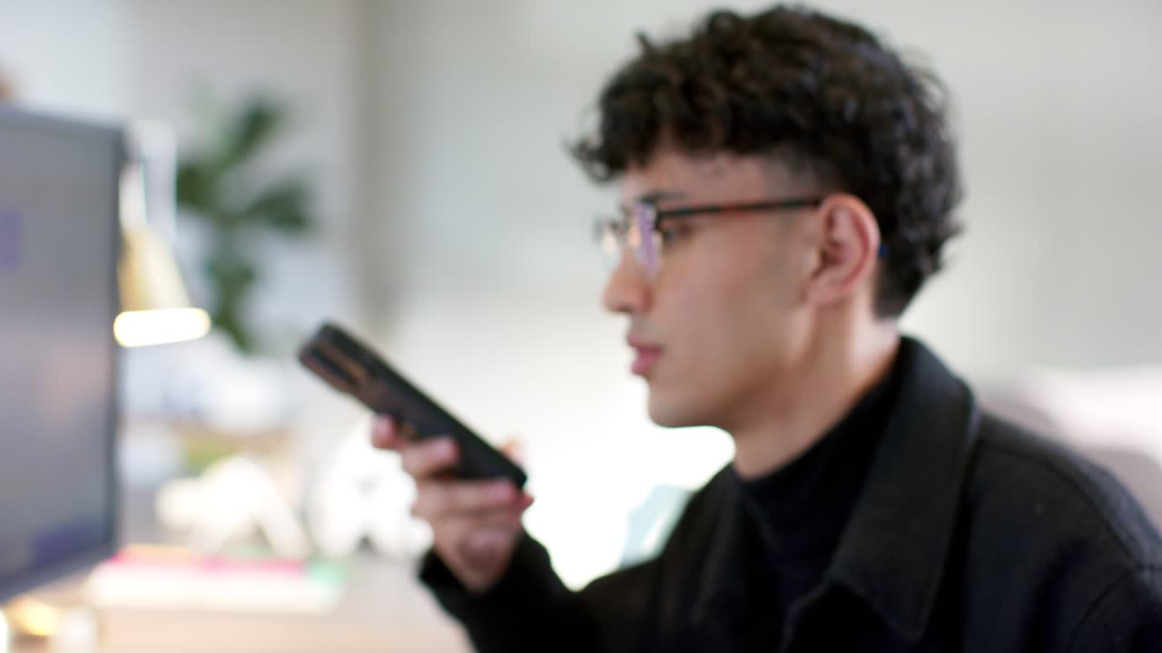 スマートフォンを見て録画を開始し、デスクでモニターを確認しながら話すアジア人男性。