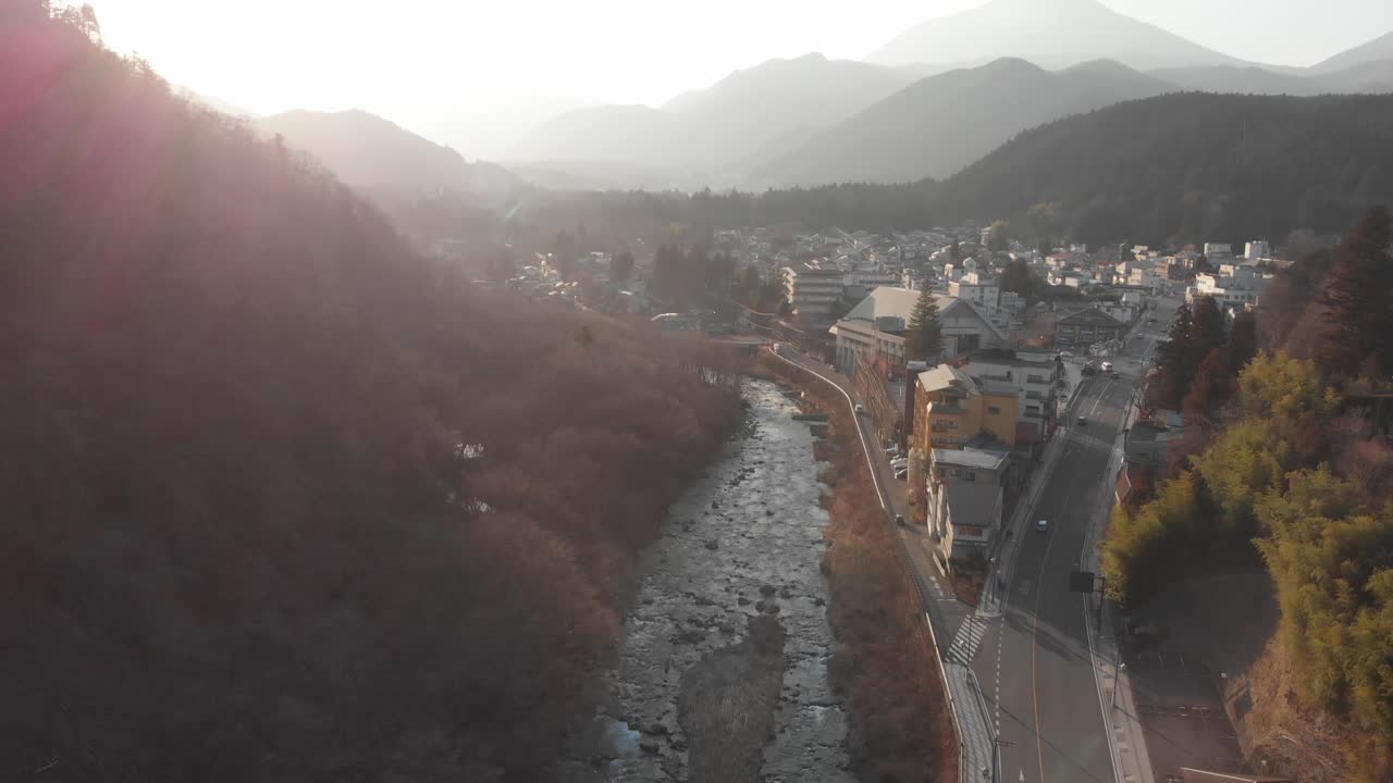 vuelo de drones, sobre la pequeña ciudad japonesa de nikko, fondo brumoso durante la hora dorada con rayos que brillan mostrando siluetas de montañas en el fondo y un río que corre por el medio