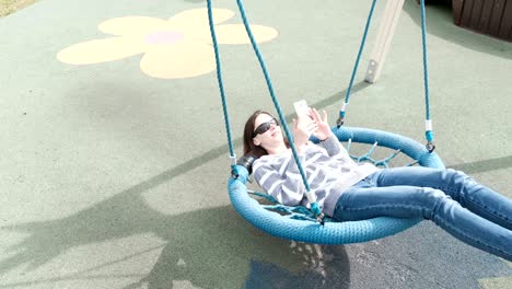 woman is swinging on empty webbed swing and talking video chat on mobile phone.