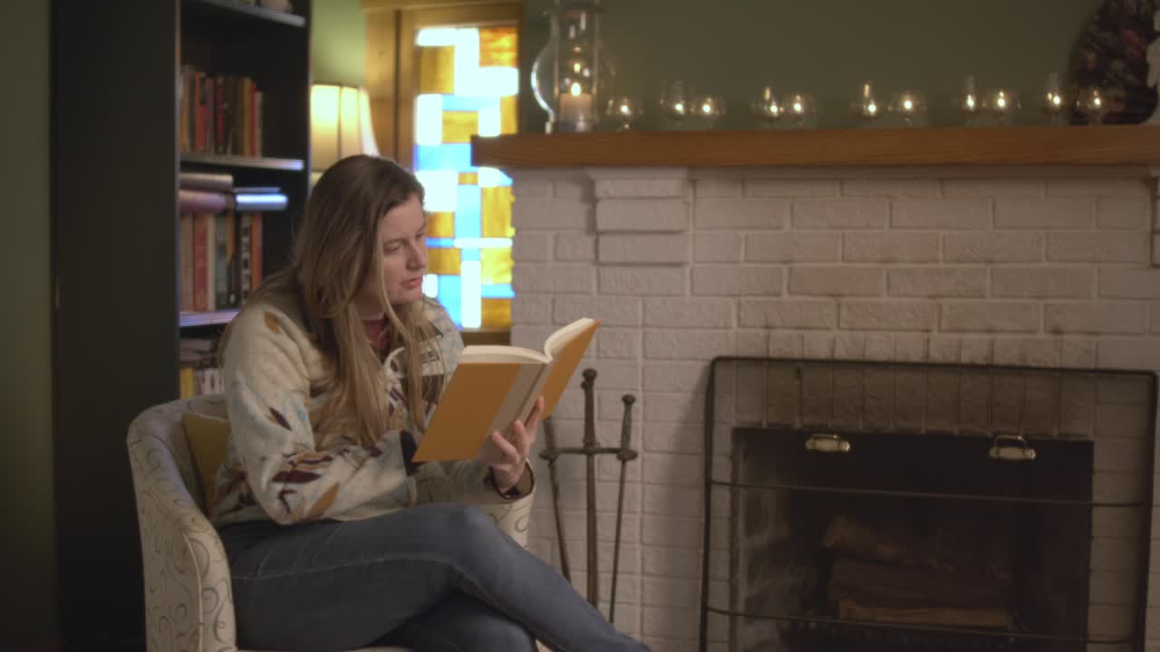 A woman intently reading a book and shocked at what she reads. Putting the book down. She reads in front of a fireplace in a cozy living room. Shot in 60 frames for slow motion