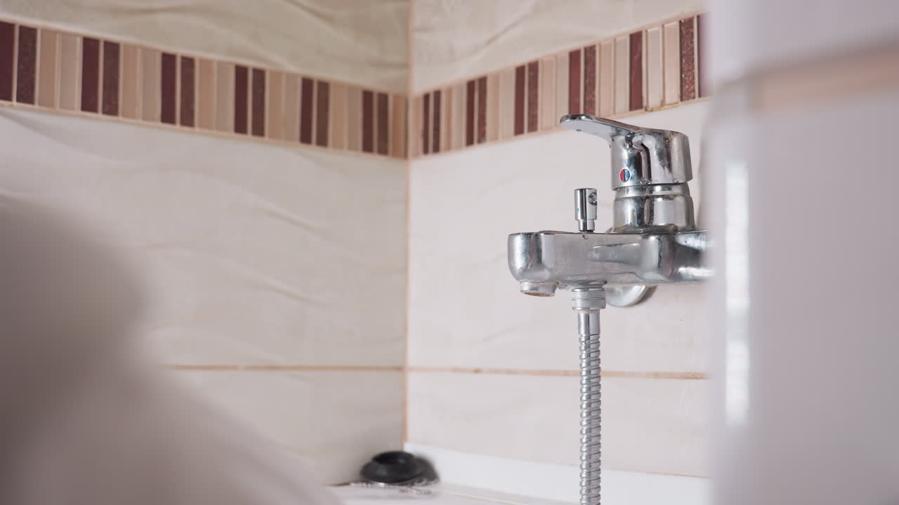 Lower view shows female hand reaching mixer tap on tiled bathroom wall, lever pushed to start water, chrome body with hose below, soft light across tiles, calm routine moment before washing