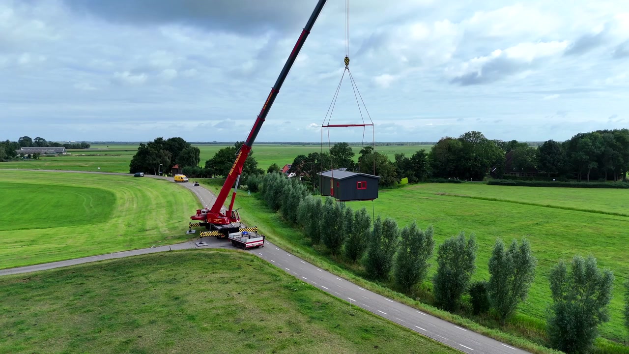 Droneoptagelse af en stor rød mobilkran parkeret på en landlig landevej, flankeret af grønne marker og enge, med en lille flok køer, der græsser i nærheden. Løfter det lille hus