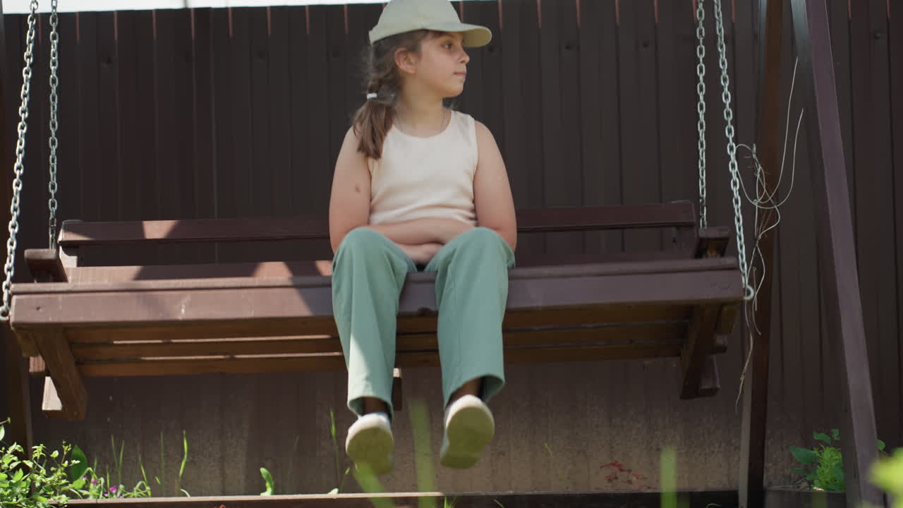 Youthful Girl Rests Calmly In Garden, Child Seated Serenely On Swing In Sunny Suburban Yard, Young Girl Patiently Swings On Bench Beneath Bright Summer Sunlight In Neighborhood Garden