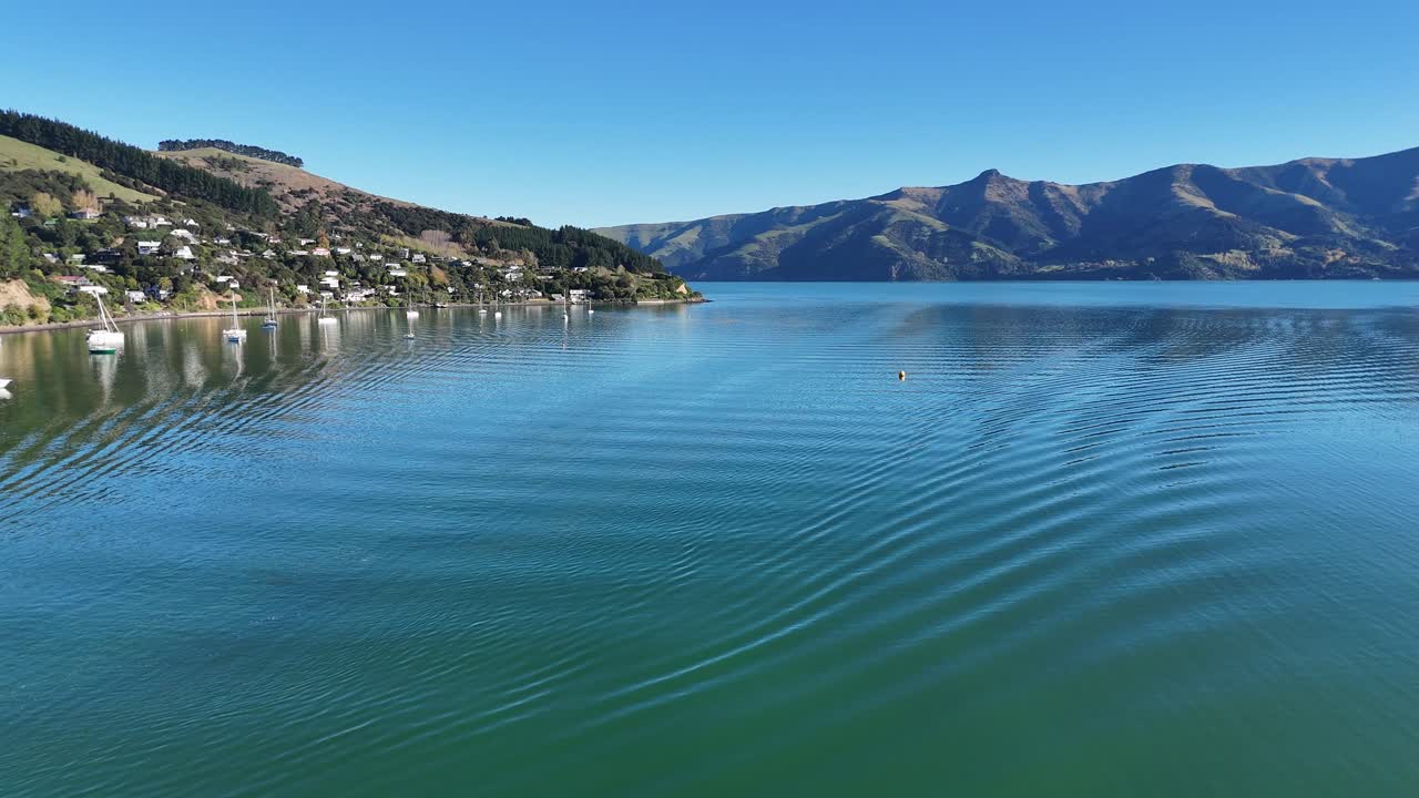 Drone captures tranquil Akaroa Bay with rippling water and distant hills under clear blue skies