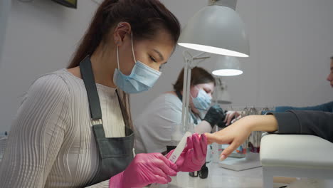 two nail technicians wearing protective masks and gloves attentively perform manicure services for clients under bright salon lamps, with one filing nails and the other applying product