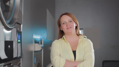 portrait of controller woman posing beside industrial washer inside modern laundromat, stainless machines glowing panels in background, confident staff in yellow shirt stands relaxed
