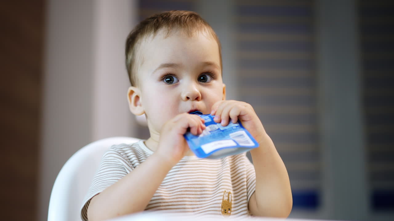 Beautiful toddler consuming factory produced fruit puree. Baby boy eats organic food from doy pack. Close up. Blurred backdrop.