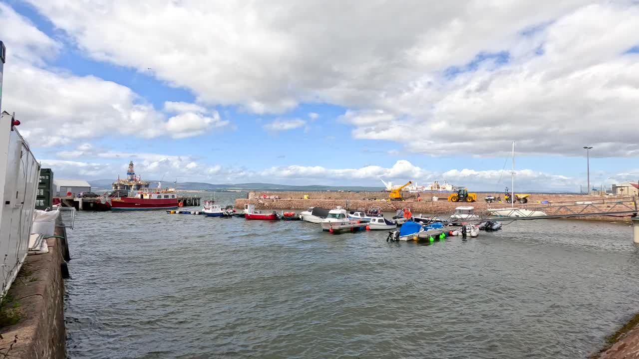 Wide shot of small boats moving into Cromarty harbor, with colorful waterfront houses and dramatic clouds. Natural daylight, steady camera, calm maritime atmosphere