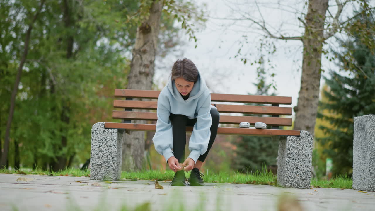 Lady sitting on concrete bench tying shoelace in outdoor park, blurred background of green trees and park elements, focused on feet and shoelaces, calm and relaxed atmosphere in nature