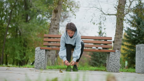 lady sitting on concrete bench tying shoelace in outdoor park, blurred background of green trees and park elements, focused on feet and shoelaces, calm and relaxed atmosphere in nature
