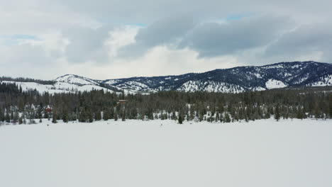 Aerial-moving-towards-frozen-lake-shore-with-trees-and-snowy-mountains