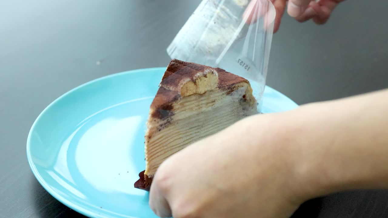 Hand carefully peels plastic wrap from tiramisu cake slice on blue plate, natural lighting