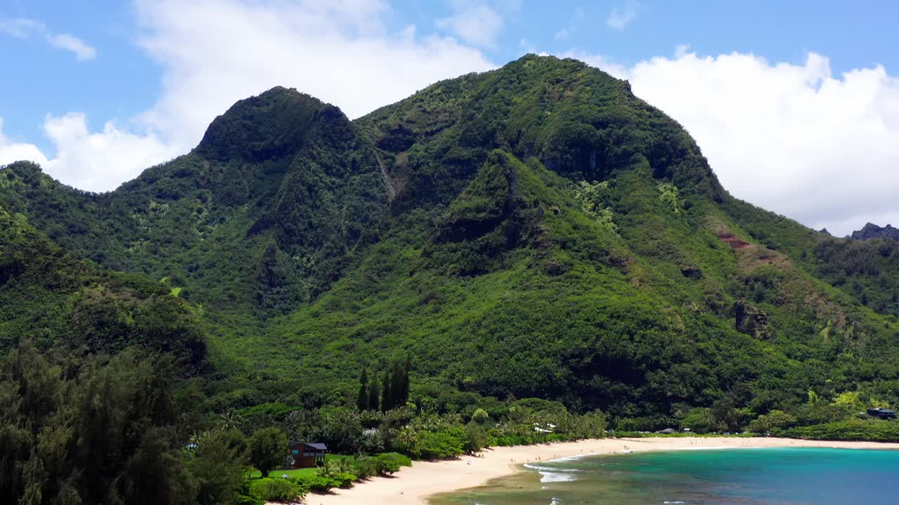 Smooth 4K aerial of Tunnels Beach in Kauai, Hawaii. Clear ocean water reveals reef patterns with snorkelers below. Lush rainforest mountains complete this tropical, cinematic view