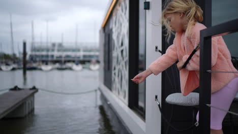 young girl in a pink sweater leans over a railing by the water, playfully tossing food into the water, surrounded by boats and a cloudy sky, capturing a joyful moment of connection with nature