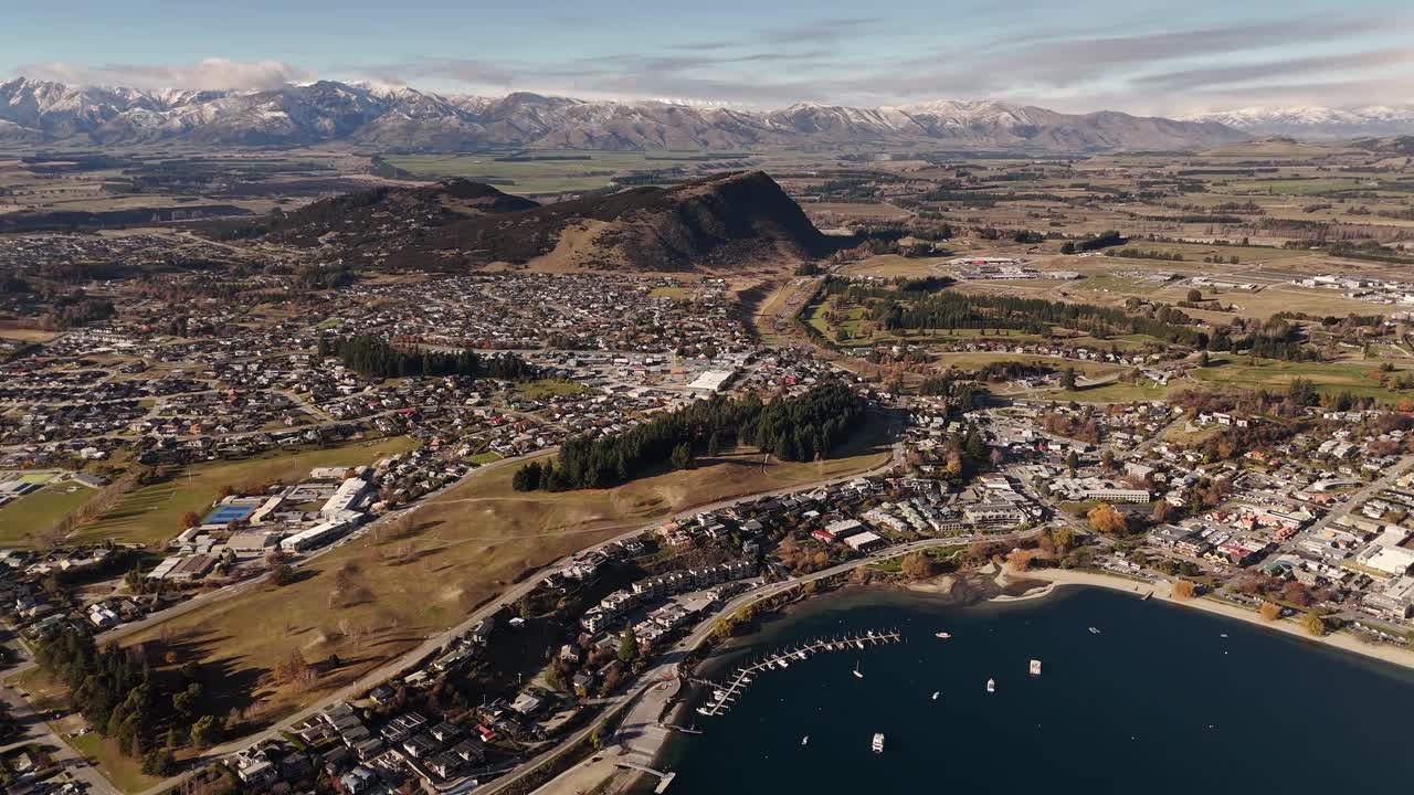 Aerial flying backwards revealing Wanaka town on lakefront with snow-capped Southern Alps mountain range in background, New Zealand