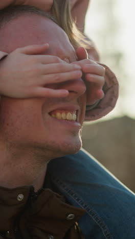 father and daughter playing in the park at sunset, with the daughter sitting on his shoulders, covering his eyes while both laugh joyfully