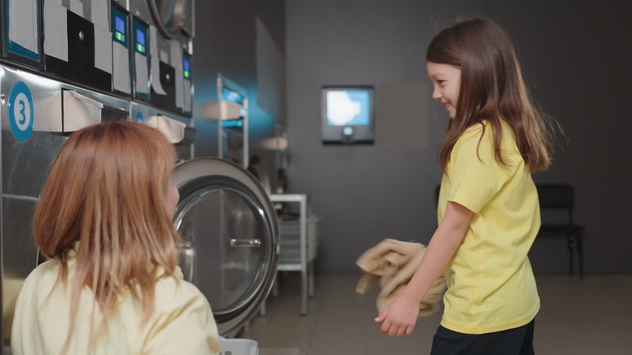 Overhead view of nurturing woman seated by washer loading clothes, little girl swings towel playfully before keeping items, teamwork mood in modern laundromat, candid family work moment