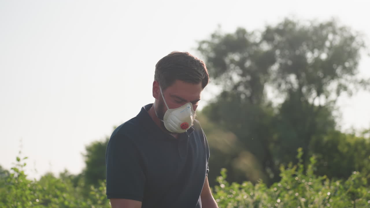 Gardener wearing protective face mask and blue gloves bends forward while preparing to fumigate plants in lush green outdoor area, with sun shining through trees in background