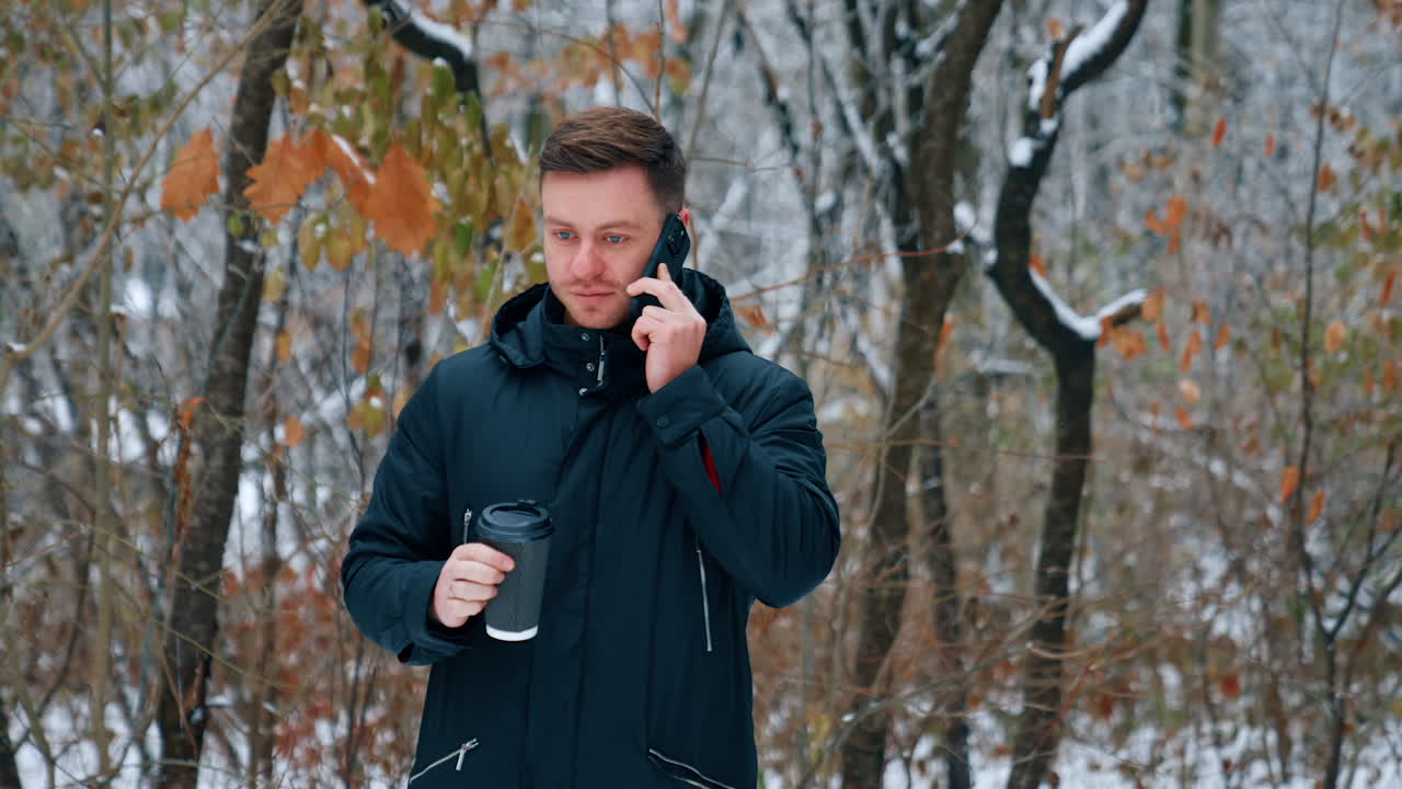 Dark-haired Caucasian male drinks coffee and speaks on the phone. Man on the walk in winter park.