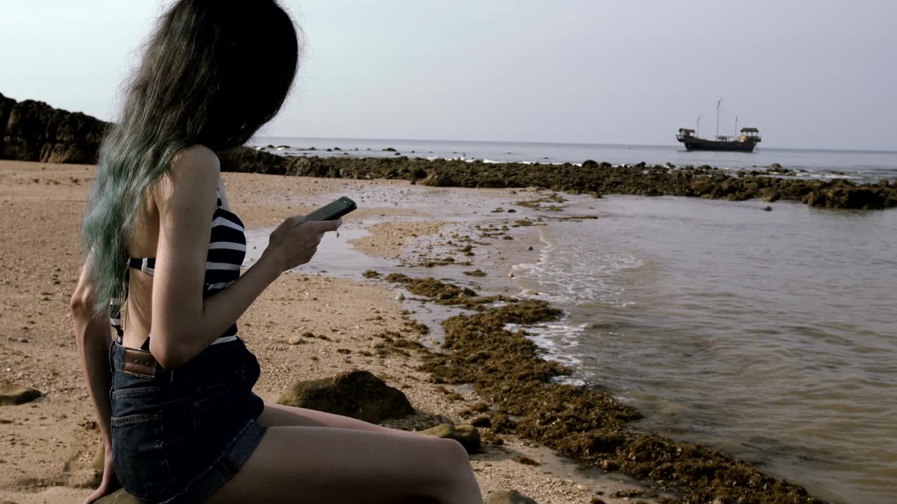 아름다운 배와 함께 바다 근처의 해변에서 휴대전화를 사용하는 젊은 관광 소녀