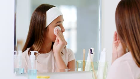 beauty,-hygiene-and-people-concept--teenage-girl-looking-in-mirror-and-cleaning-face-skin-with-cotton-disc-at-bathroom