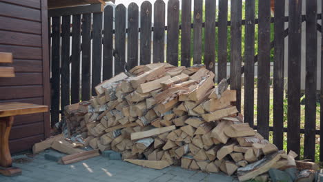 stacked birch logs arranged neatly against dark wooden fence on paved ground, sunlight filtering through surrounding trees, rustic outdoor woodpile for cottage fireplace, textured forest ambiance