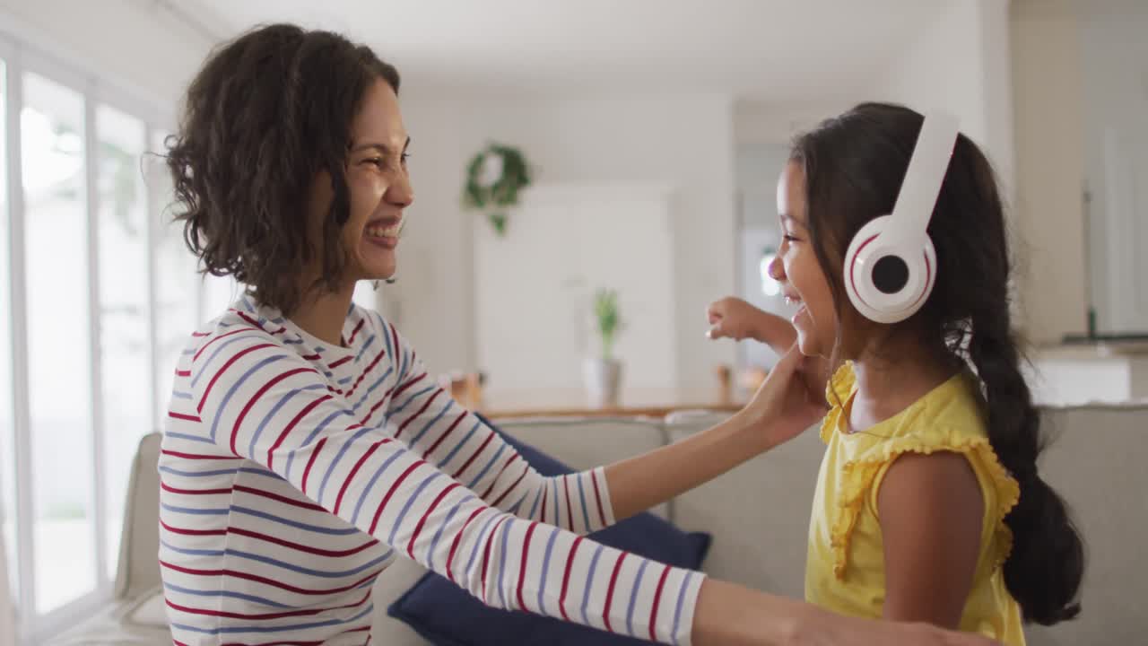 Happy hispanic mother and daughter wearing headphones listening to music sitting on sofa