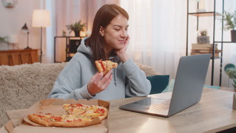 Happy woman eating pizza sitting on sofa at table watching movie or series online on laptop at home