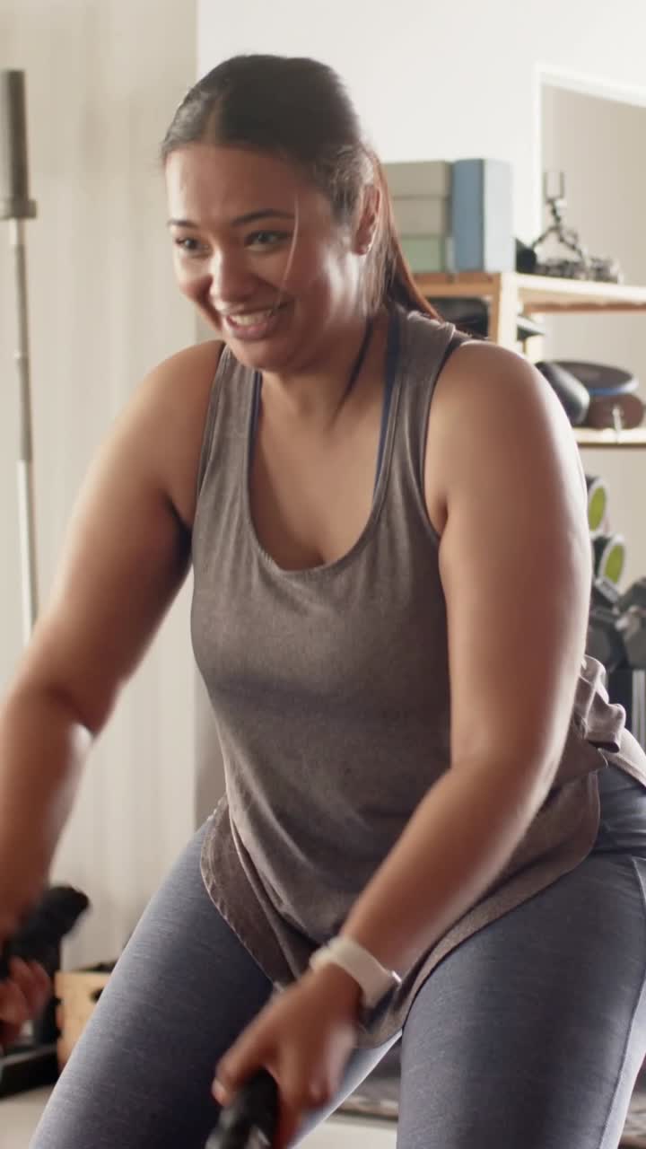 Video verticale: Afferrando la battle rope, una donna esegue tirate alternate nella palestra di casa, in canotta per il cardio.