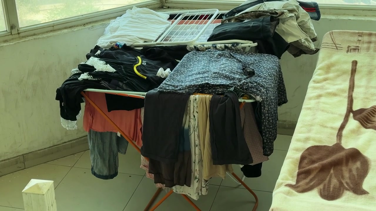 A static shot of an indoor laundry drying rack full of colorful washed clothes. The folding rack sits next to a bed by large windows overlooking an urban street in bright daytime sunlight.