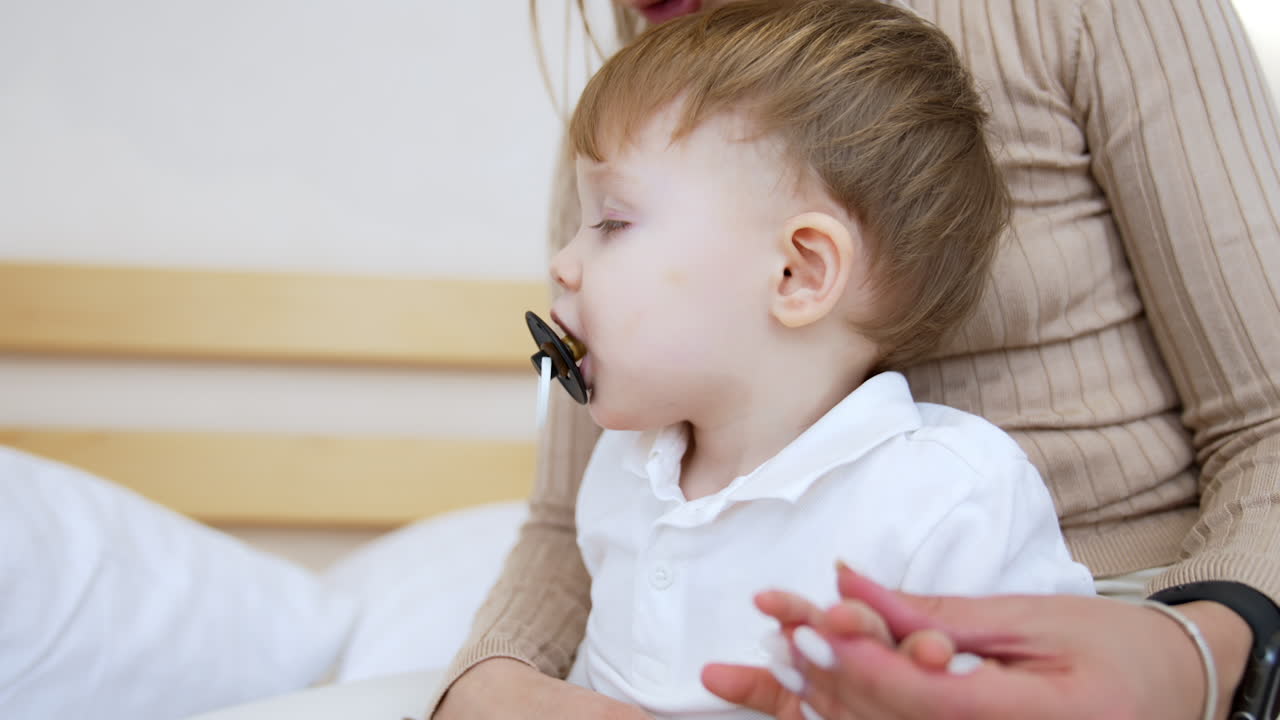 Cute baby boy with pacifier in mouth sits in mom's hands. Unrecognized mother stroking her child's tiny fingers. Close up.