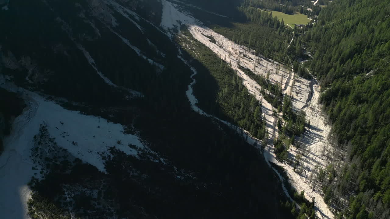Ranní letecký přelet nad suchým říčním korytem poblíž Lago di Braies v italských Alpách. Vycházející slunce vrhá dlouhé stíny přes horské údolí a lesy. Natočeno ve 4K, Apple ProRes při 25 snímcích za sekundu.