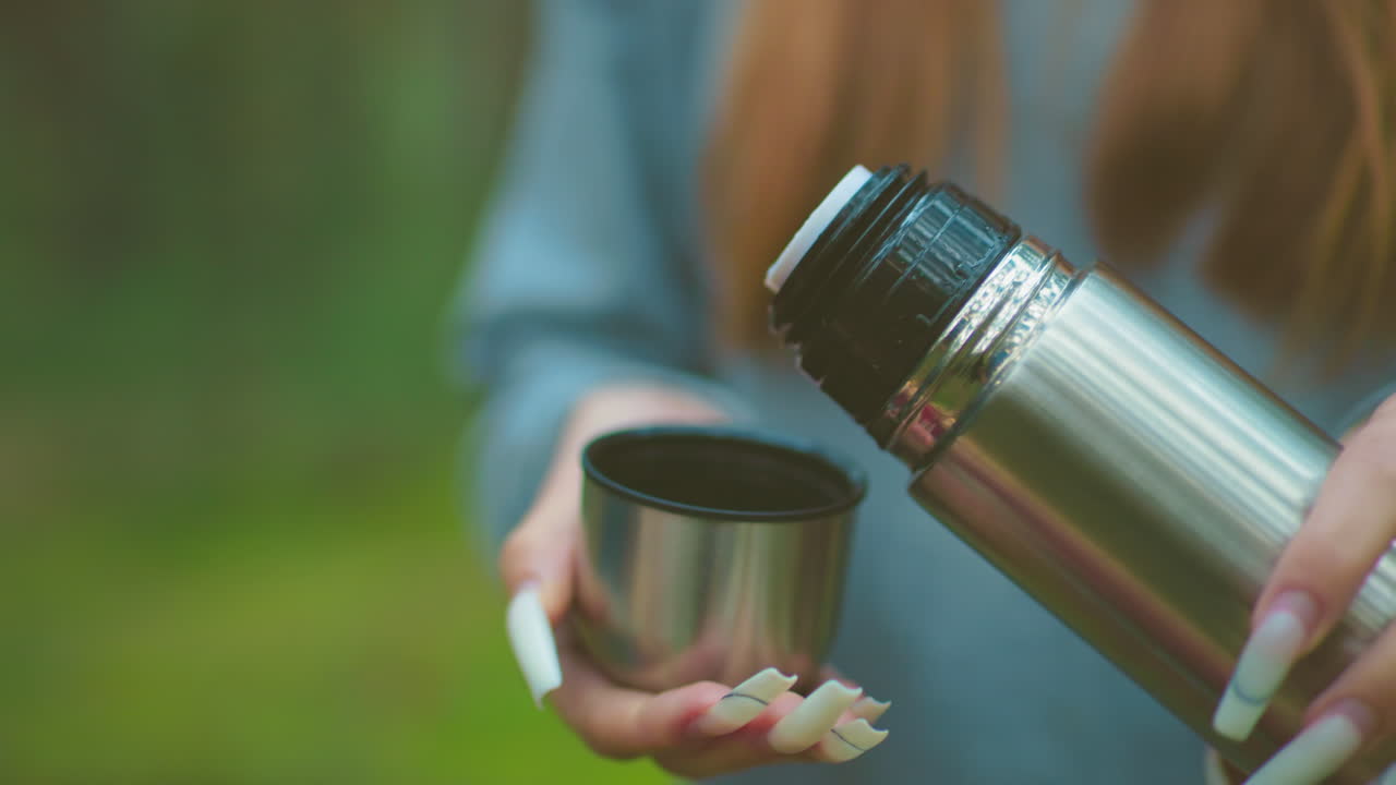 primer plano de alguien con uñas bien pulidas presionando la cubierta del termo para verter agua en la tapa, contra un fondo verde borroso, destacando el manejo cuidadoso de la bebida al aire libre