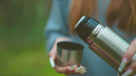Close-up of someone with well polished nails pressing thermos cover to pour water into lid, against blurred greenery background, highlighting careful handling of drink outdoors