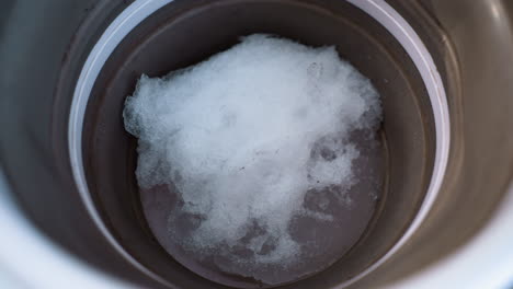 high angle close up of brown bucket containing melting snow placed on clear plastic table under soft light, with water droplets and blurred background creating calm, minimalistic winter feel indoors