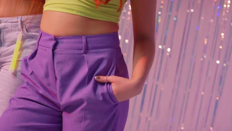 studio close up of two women showing hips and torsos standing in front of tinsel curtain against pink background