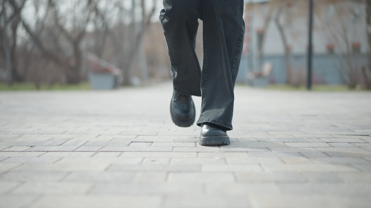 Low-angle view of woman s feet in black leather boots and dark pants walking across paved park path during winter day, capturing motion, stride, seasonal attire, and casual street style in soft daylight