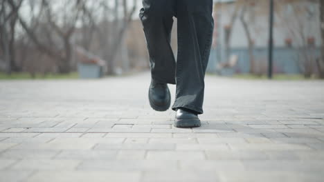 Low-angle view of woman s feet in black leather boots and dark pants walking across paved park path during winter day, capturing motion, stride, seasonal attire, and casual street style in soft daylight