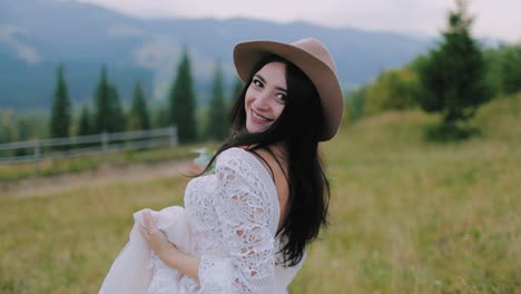dark-haired bride walking happily in nature. backside view of a pretty woman in a wedding dress and hat on mountains background.