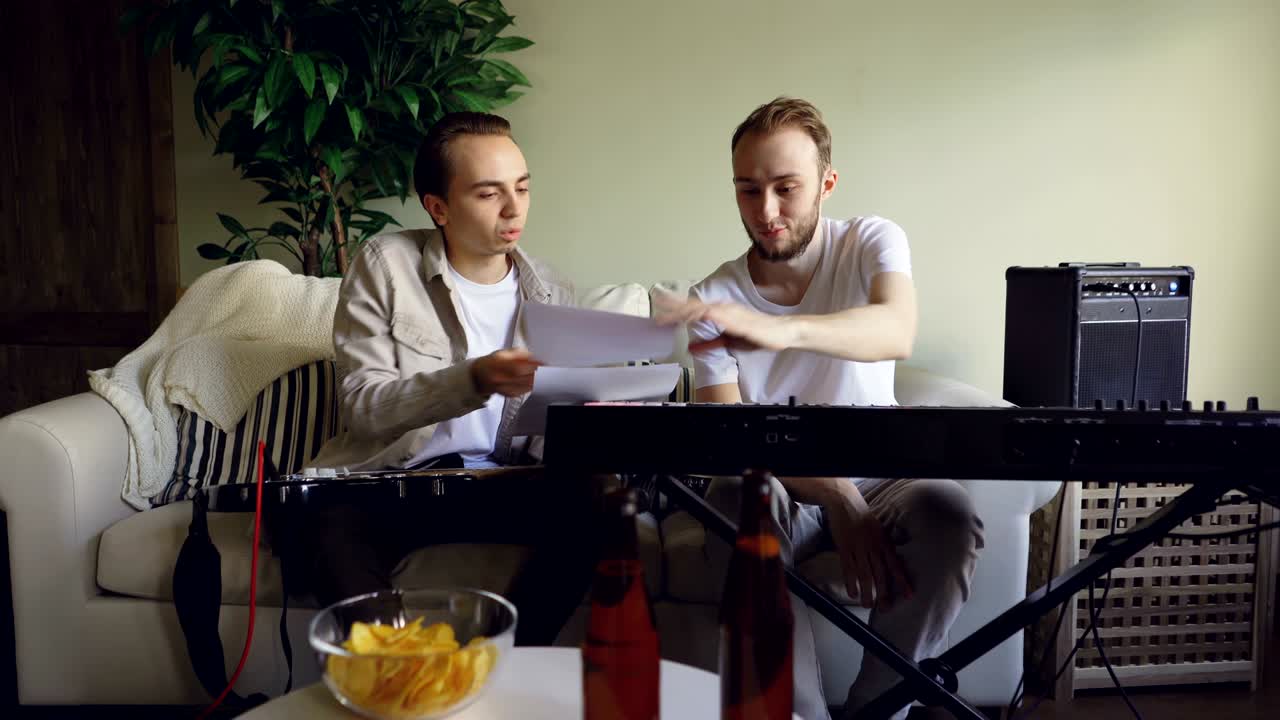 гитарист и клавишник разговаривают, глядя на музыкальный лист и обсуждая тексты во время репетиционного перерыва. электрические музыкальные инструменты, напитки и закуски видны.