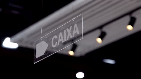 transparent overhead sign with word 'caixa' under ceiling lights in retail environment
