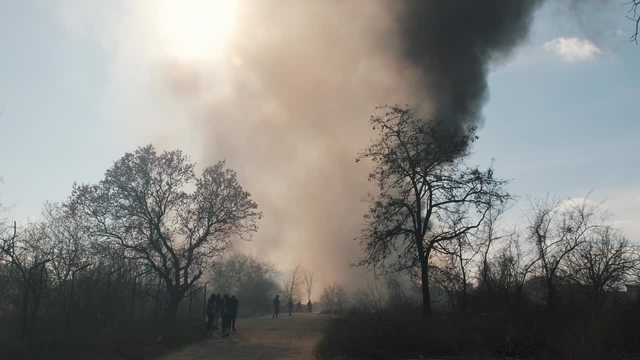 High column thick smoke and fire in city park against blue sky, bright sun disk, sun rays slide forward , arson against of trees in spring lens flare. Protest, criminal incident. Flames. Slow motion